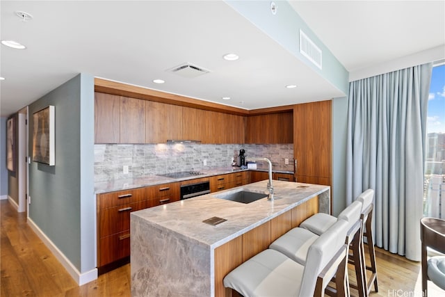 kitchen featuring backsplash, stainless steel oven, a kitchen island with sink, light hardwood / wood-style floors, and sink