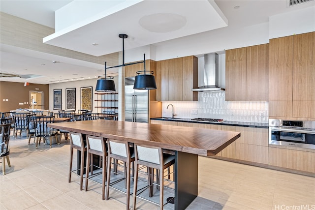 kitchen featuring sink, a kitchen breakfast bar, stainless steel appliances, decorative backsplash, and wall chimney range hood