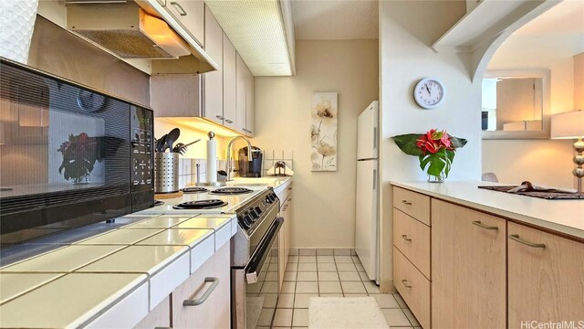 kitchen with stainless steel electric range, tile counters, light tile patterned floors, sink, and light brown cabinetry