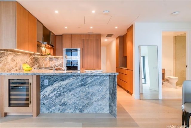 kitchen featuring tasteful backsplash, stainless steel double oven, light stone counters, light wood-type flooring, and beverage cooler