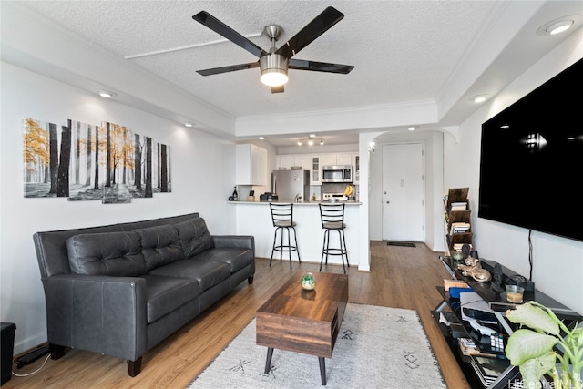 living room featuring crown molding, light wood-type flooring, a textured ceiling, a raised ceiling, and ceiling fan