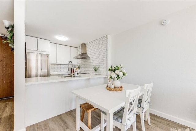 kitchen with stainless steel fridge, wall chimney exhaust hood, a peninsula, light countertops, and white cabinetry