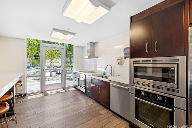 kitchen featuring sink, stainless steel appliances, wall chimney range hood, light hardwood / wood-style floors, and dark brown cabinets