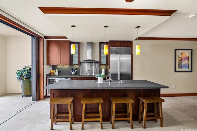 kitchen with a breakfast bar, dark countertops, wall chimney range hood, a center island with sink, and built in fridge