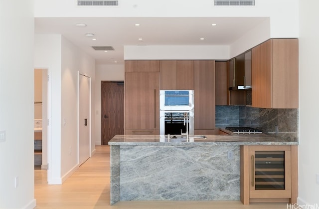 kitchen featuring wall chimney exhaust hood, wine cooler, kitchen peninsula, stone countertops, and light wood-type flooring