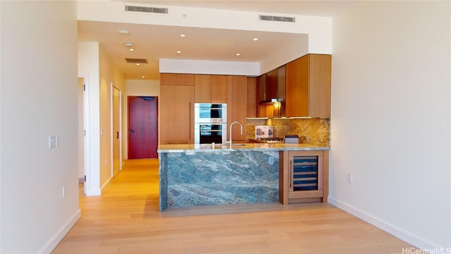 kitchen featuring stainless steel double oven, wine cooler, light stone counters, backsplash, and light hardwood / wood-style floors