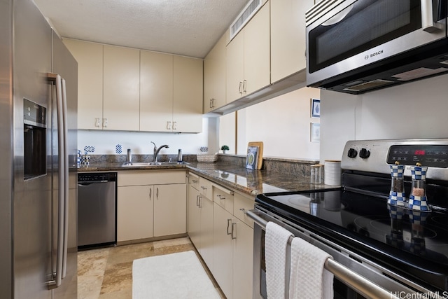 kitchen with appliances with stainless steel finishes, dark stone counters, a textured ceiling, sink, and cream cabinetry