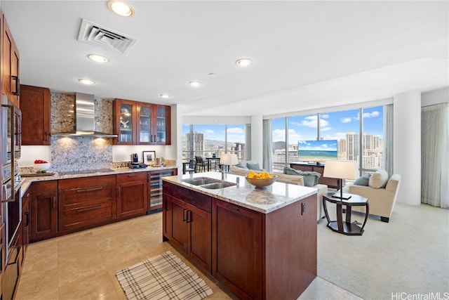 kitchen with light stone countertops, backsplash, wall chimney range hood, wine cooler, and an island with sink