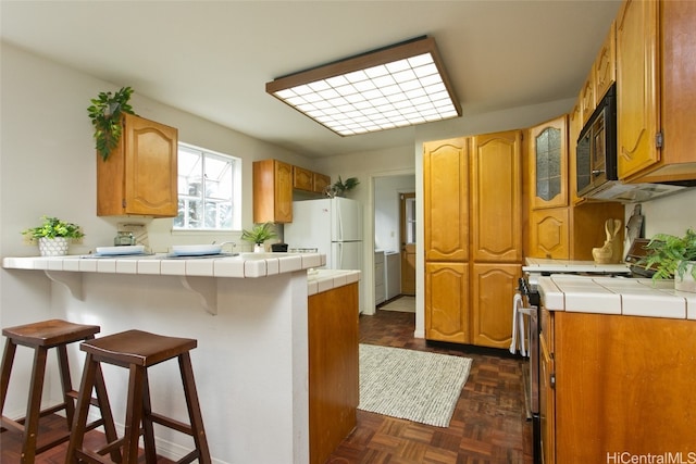 kitchen with kitchen peninsula, a kitchen bar, white appliances, dark parquet floors, and tile countertops