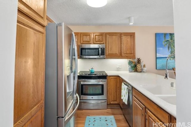 kitchen with light wood finished floors, appliances with stainless steel finishes, brown cabinetry, a sink, and a textured ceiling