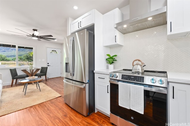 kitchen with stainless steel appliances, ceiling fan, wall chimney range hood, light hardwood / wood-style floors, and white cabinetry