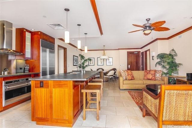 kitchen with a kitchen island with sink, wall chimney exhaust hood, ornamental molding, appliances with stainless steel finishes, and decorative light fixtures