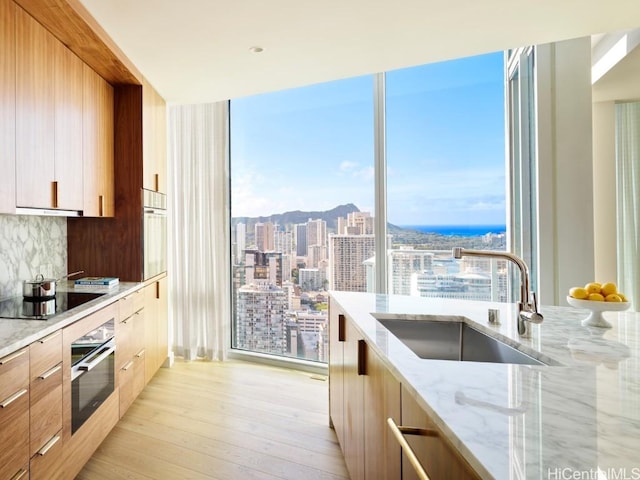 kitchen with stainless steel oven, sink, tasteful backsplash, light stone counters, and light wood-type flooring