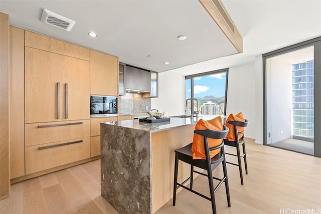 kitchen with built in microwave, light brown cabinets, a kitchen breakfast bar, light hardwood / wood-style flooring, and a kitchen island with sink