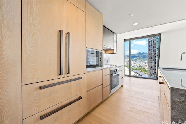 kitchen with light brown cabinetry, black microwave, sink, light hardwood / wood-style floors, and oven