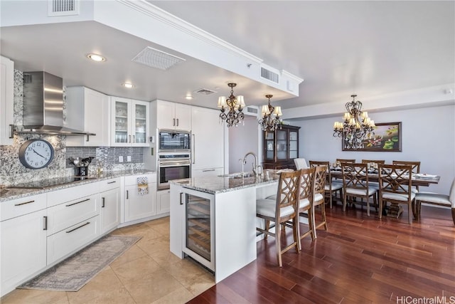 kitchen featuring a kitchen island with sink, white cabinets, wall chimney range hood, appliances with stainless steel finishes, and beverage cooler