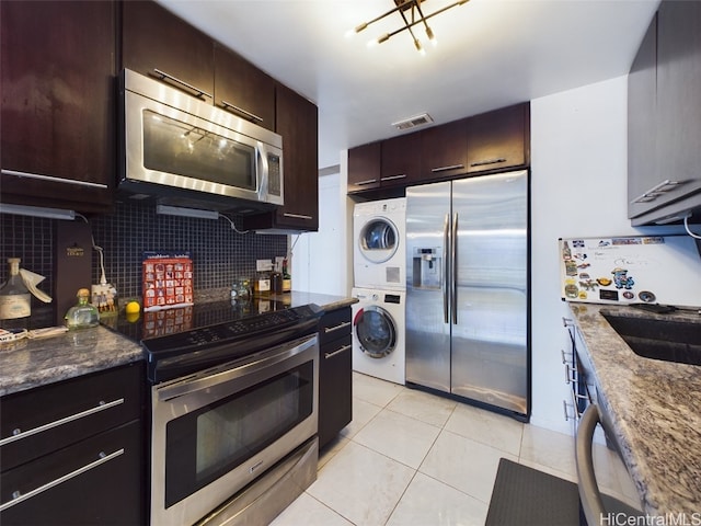 kitchen featuring appliances with stainless steel finishes, backsplash, dark stone counters, light tile patterned floors, and stacked washer / dryer