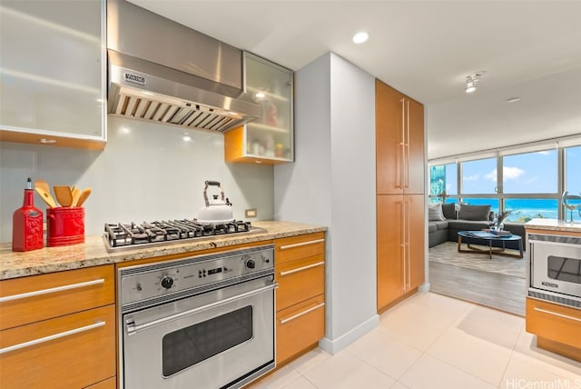 kitchen featuring wall chimney exhaust hood, stainless steel appliances, light tile patterned floors, light stone counters, and a water view