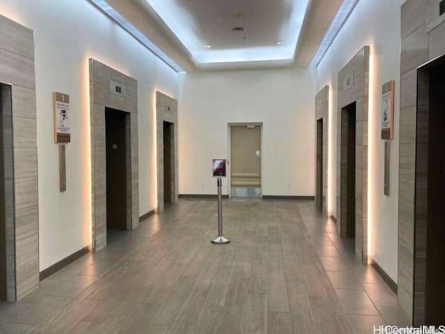 corridor with elevator, baseboards, a tray ceiling, and wood finished floors