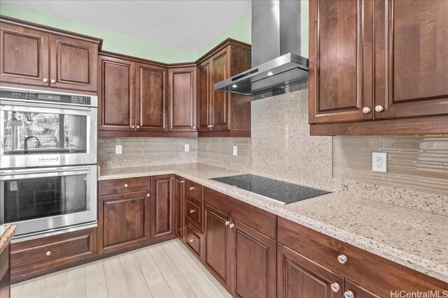 kitchen with stainless steel double oven, wall chimney range hood, light stone counters, backsplash, and black electric stovetop