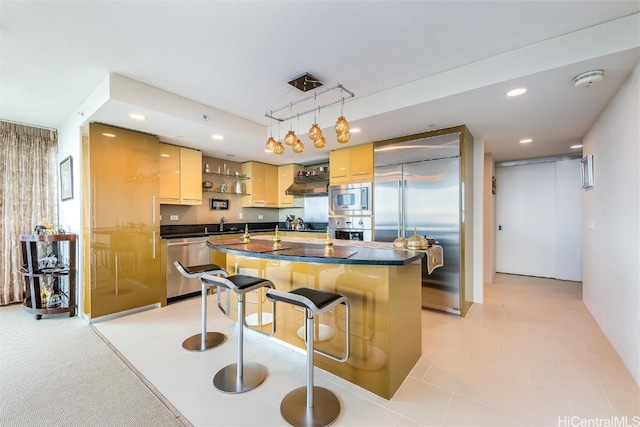 kitchen with a breakfast bar, a kitchen island with sink, hanging light fixtures, built in appliances, and light brown cabinetry