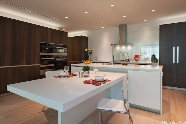 kitchen featuring dark brown cabinetry, light wood-type flooring, a kitchen island with sink, a breakfast bar area, and wall chimney exhaust hood