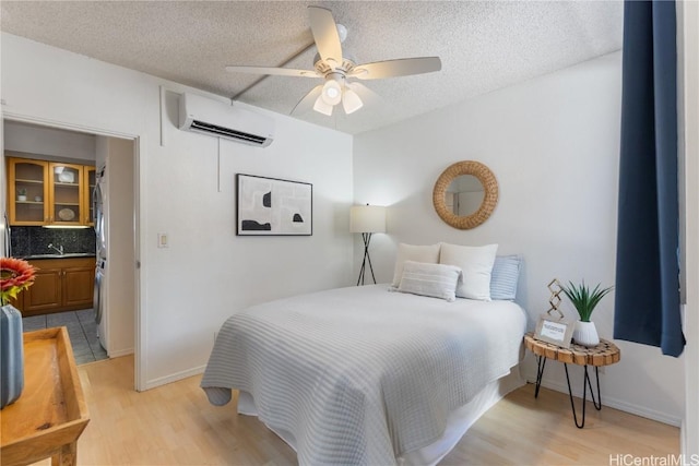 bedroom featuring a textured ceiling, light wood-style flooring, a ceiling fan, baseboards, and a wall mounted AC