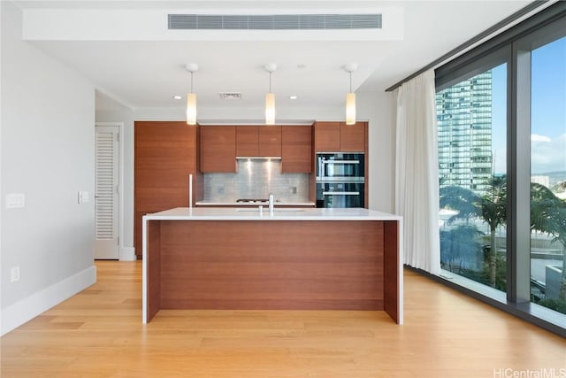 kitchen with tasteful backsplash, black double oven, a center island with sink, light hardwood / wood-style floors, and hanging light fixtures