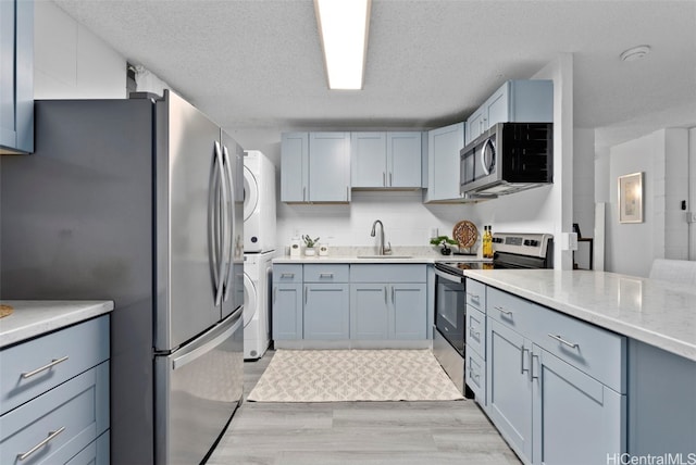 kitchen with appliances with stainless steel finishes, sink, stacked washer and clothes dryer, a textured ceiling, and light hardwood / wood-style flooring