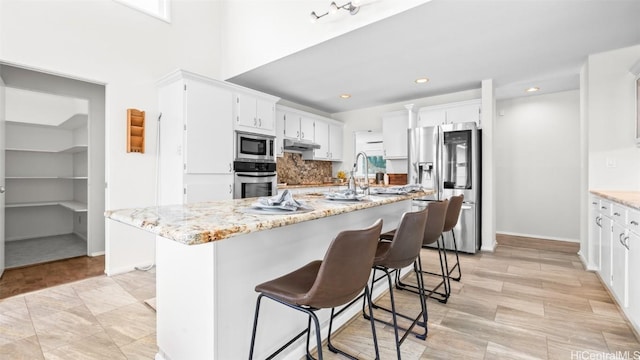 kitchen with under cabinet range hood, light stone counters, appliances with stainless steel finishes, and white cabinets