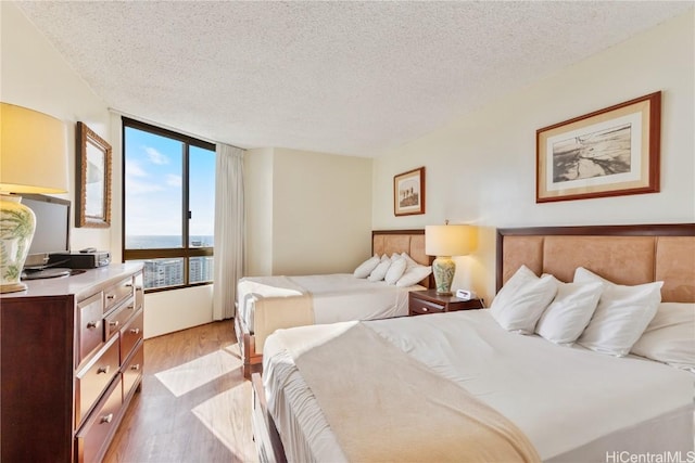 bedroom featuring a water view, light wood-type flooring, floor to ceiling windows, and a textured ceiling