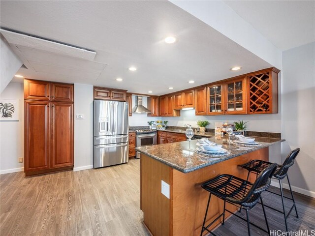 kitchen featuring kitchen peninsula, wall chimney exhaust hood, light wood-type flooring, appliances with stainless steel finishes, and dark stone countertops