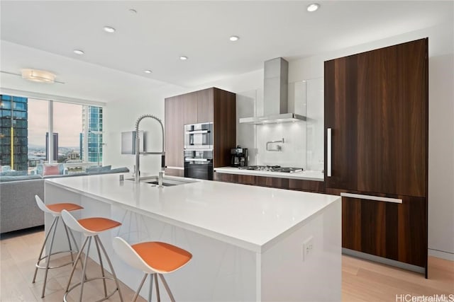 kitchen with stainless steel appliances, an island with sink, and wall chimney range hood