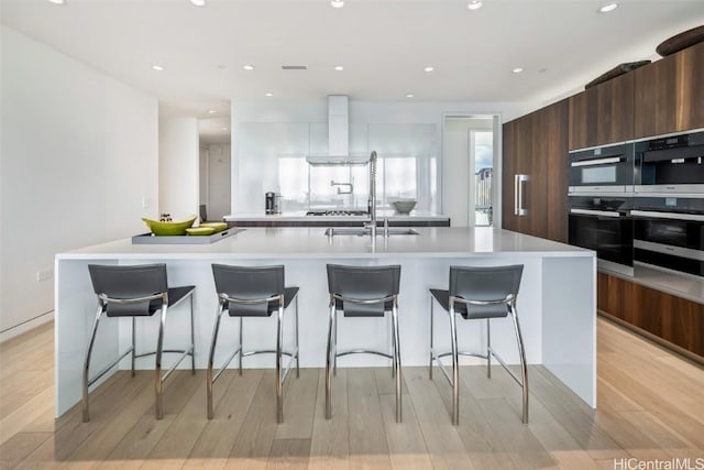 kitchen with a kitchen breakfast bar, a large island with sink, stainless steel double oven, and wall chimney range hood