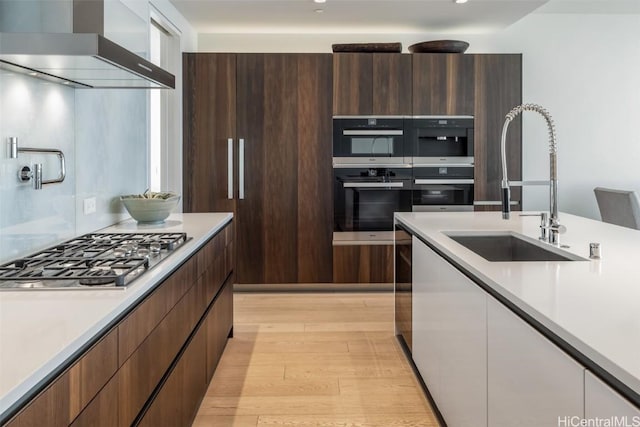 kitchen with stainless steel gas stovetop, wall chimney range hood, light hardwood / wood-style floors, sink, and double oven