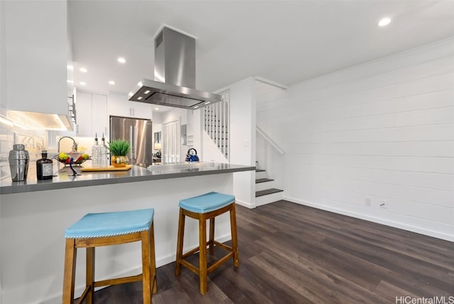 kitchen with a kitchen bar, island range hood, stainless steel refrigerator, kitchen peninsula, and white cabinets