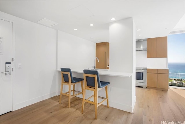 kitchen with light hardwood / wood-style flooring, wall chimney exhaust hood, a water view, stainless steel range, and a breakfast bar