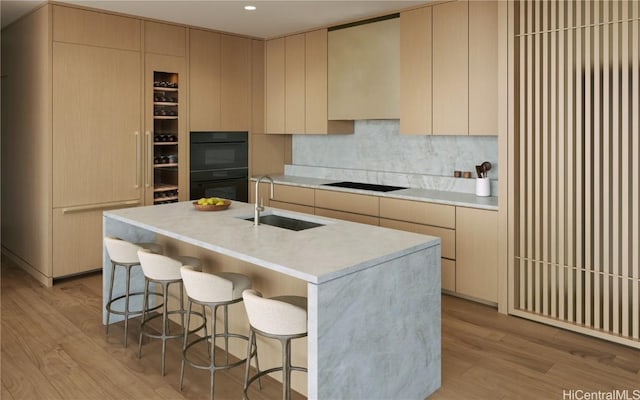 kitchen with sink, black appliances, light wood-type flooring, a kitchen island with sink, and backsplash