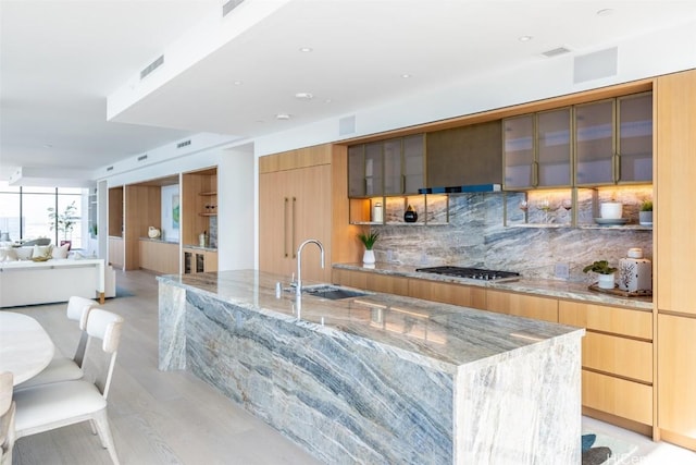 kitchen with sink, a center island with sink, stainless steel gas stovetop, light stone countertops, and decorative backsplash