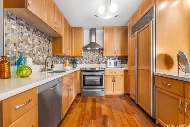 kitchen with wall chimney range hood, sink, light hardwood / wood-style flooring, appliances with stainless steel finishes, and decorative backsplash