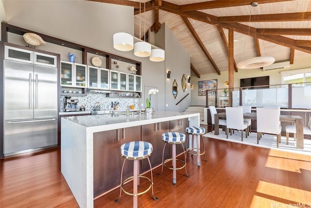 kitchen with decorative light fixtures, wood-type flooring, decorative backsplash, built in refrigerator, and a kitchen island with sink