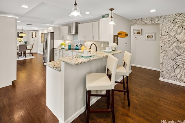 kitchen with white cabinets, a kitchen bar, kitchen peninsula, stainless steel refrigerator with ice dispenser, and wall chimney exhaust hood