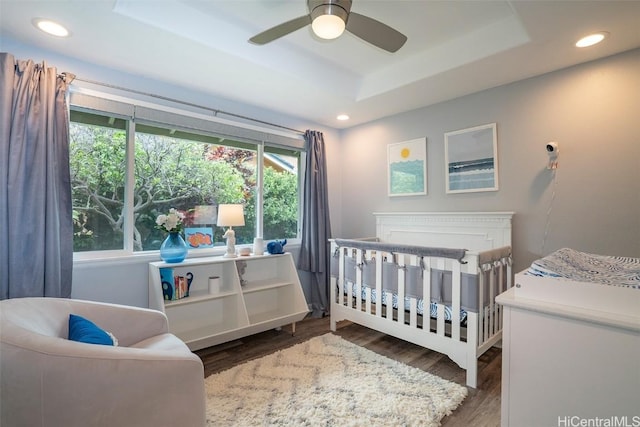 bedroom featuring dark hardwood / wood-style floors, ceiling fan, a nursery area, and a tray ceiling