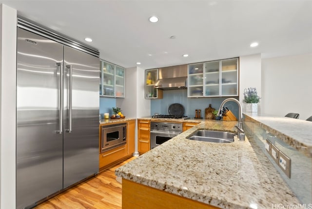 kitchen with sink, light stone counters, built in appliances, light hardwood / wood-style floors, and wall chimney range hood