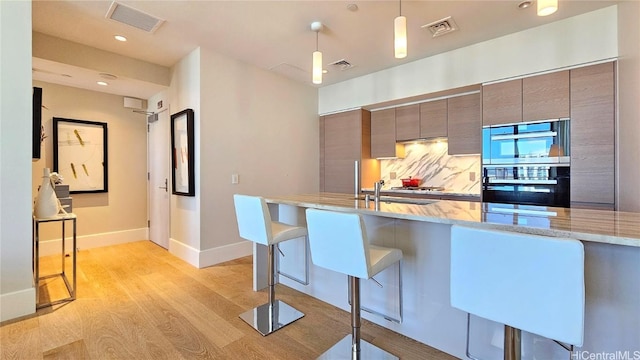 kitchen with a breakfast bar area, light wood-type flooring, double oven, pendant lighting, and light stone countertops