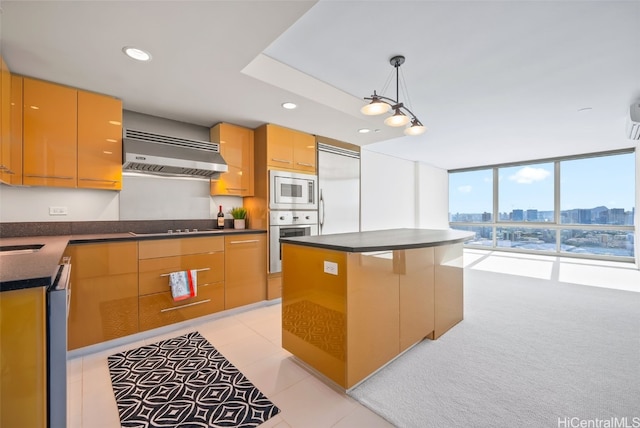 kitchen with wall chimney range hood, a wall of windows, a center island, built in appliances, and decorative light fixtures