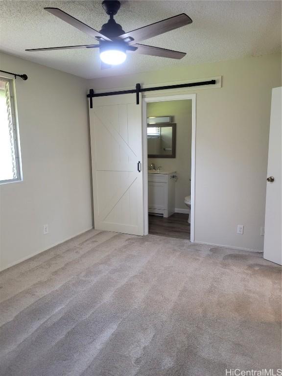 unfurnished bedroom featuring ceiling fan, ensuite bathroom, a textured ceiling, light colored carpet, and a barn door