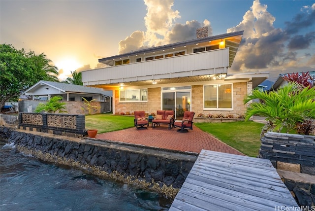 back house at dusk with an outdoor living space, a patio area, a balcony, and a lawn