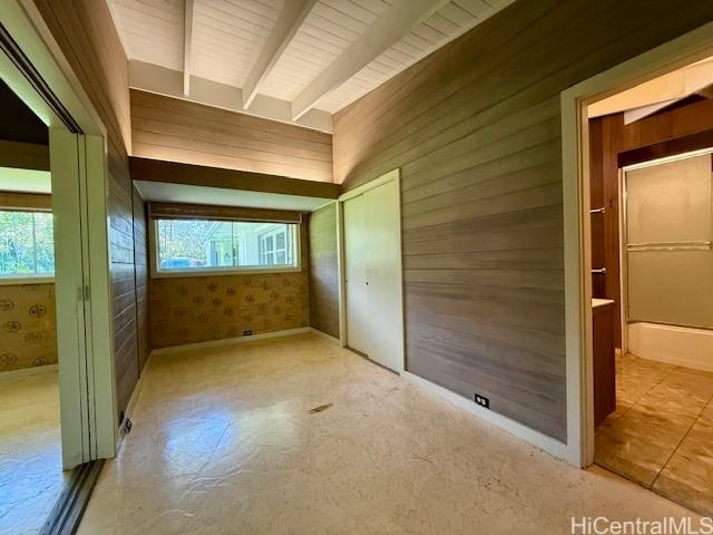 unfurnished bedroom with multiple windows, ensuite bathroom, beamed ceiling, and wood walls