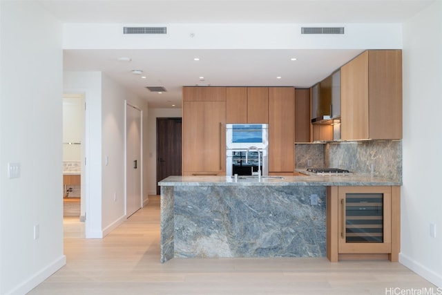 kitchen with stainless steel appliances, light stone counters, wine cooler, decorative backsplash, and wall chimney exhaust hood